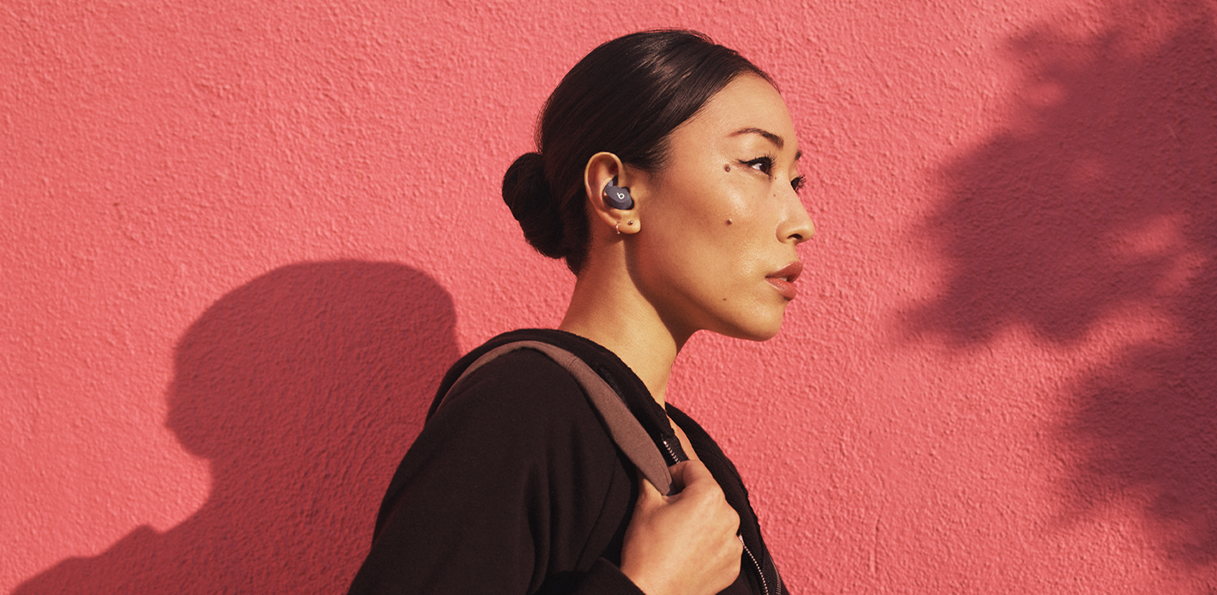 Une femme asiatique portant des Beats Fit Pro et marchant dehors devant un mur rouge